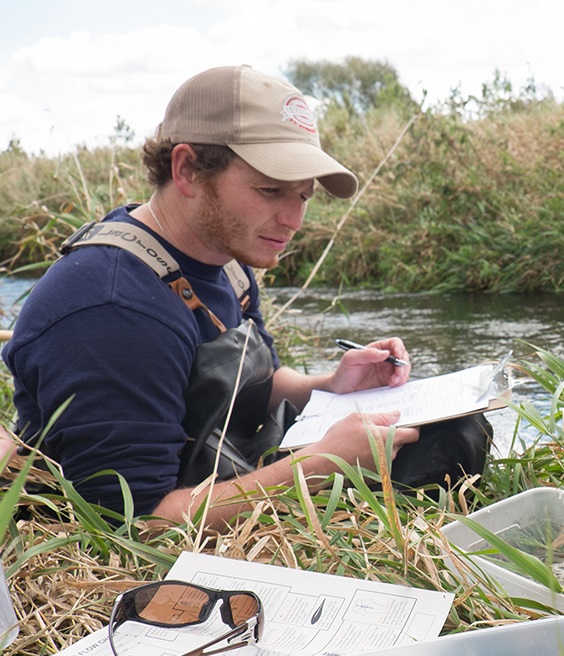 Young man wearing a ball cap sitting near a river and taking notes on a piece of paper.